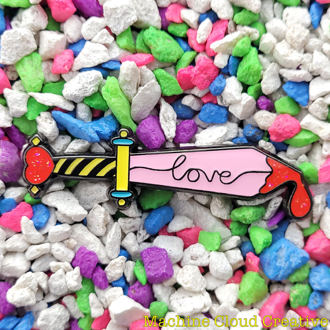 An enamel pin of a dagger with a heart on the hilt, a drop of blood on the tip, and the word love written on the fuller of the blade.