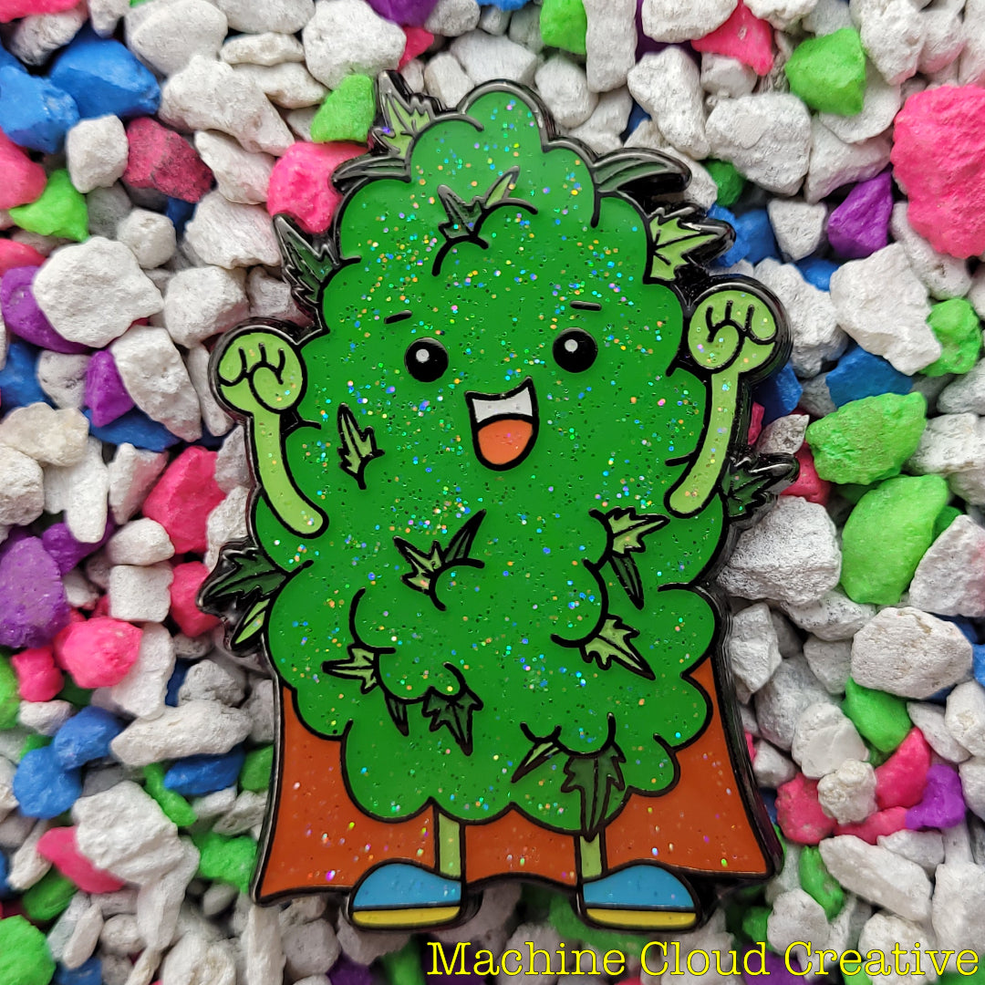 An enamel pin of a cartoon sativa cannabis nug wearing a red cape.