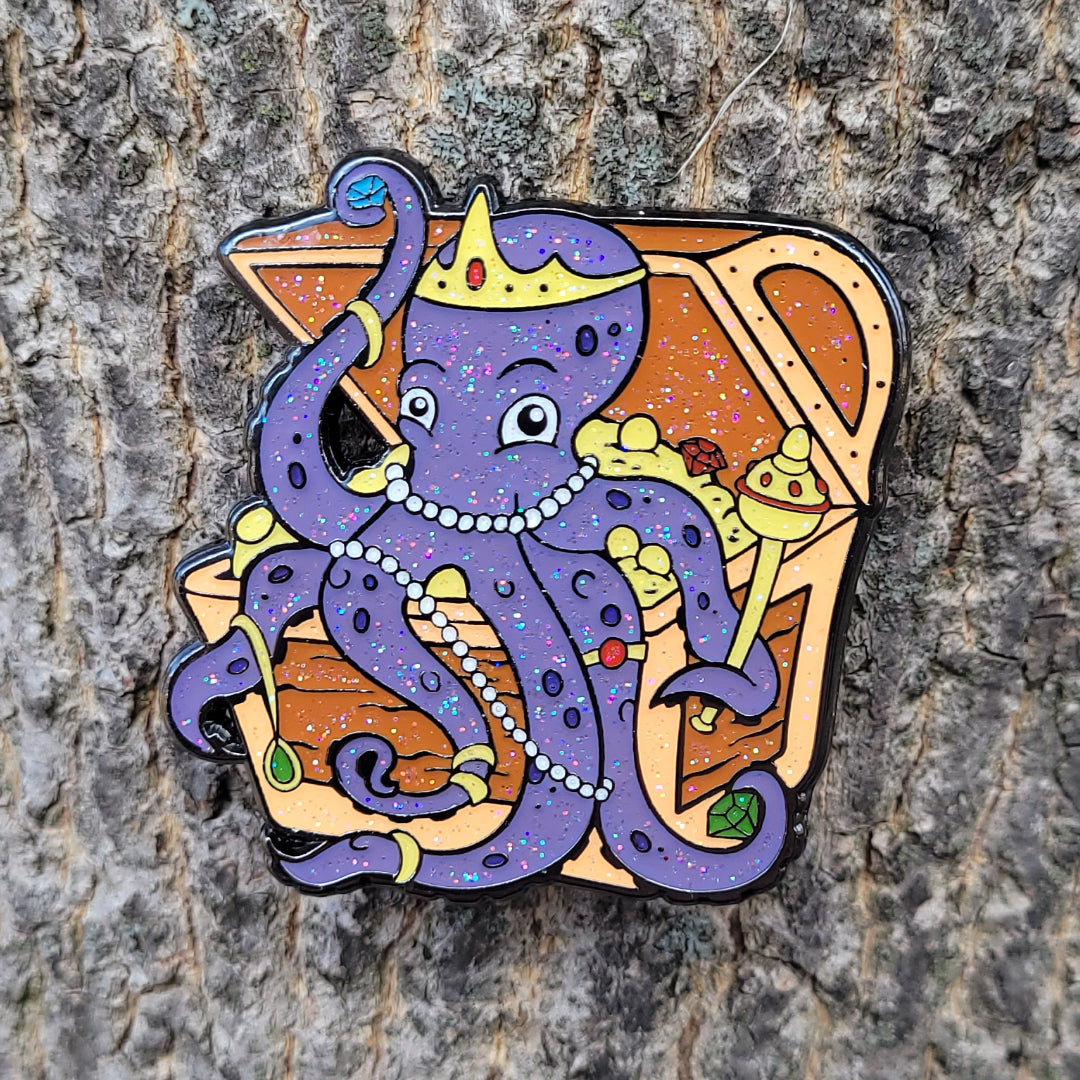 An enamel pin of an octopus sitting in a treasure chest and the pin is attached to tree bark.